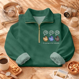 May include: A dark green quarter-zip sweatshirt with a white interior and a gold zipper. The sweatshirt features a pixelated design of three chickens and the text "I'm starting to feel exhausted..."