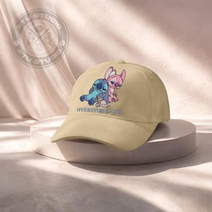 May include: A tan baseball cap featuring a blue and pink cartoon character design with the text "OVERSTIMULATED" in blue. The cap has a curved brim and a classic six-panel design.