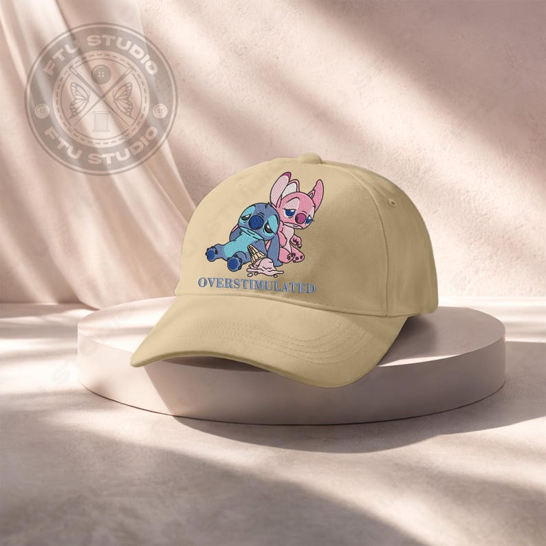 May include: A tan baseball cap featuring a blue and pink cartoon character design with the text "OVERSTIMULATED" in blue. The cap has a curved brim and a classic six-panel design.