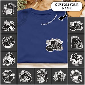 Puede incluir: Una camiseta azul marino con un gráfico de gato Druida en el bolsillo y el texto "Custom name" bordado en el cuello. La camiseta está rodeada de varias pegatinas de personajes de gatos en blanco y negro, incluyendo "Cleric", "Paladin" y "Rogue".