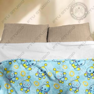 May include: A blue and yellow fleece blanket with a repeating pattern of a blue cartoon character with yellow eyes and a yellow ring. The character is on a light blue background with yellow rings.