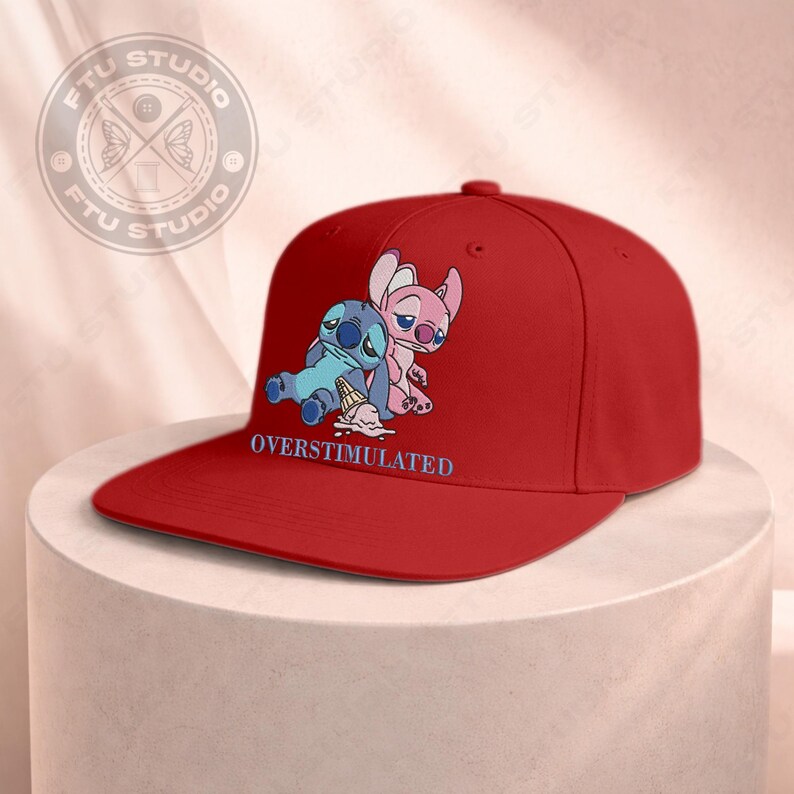 May include: A red baseball cap featuring a graphic of two cartoon characters, one blue and one pink, with the word "OVERSTIMULATED" printed below. The cap has a flat brim and a rounded crown.