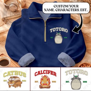 Könnte beinhalten: Ein marineblaues Fleece-Pullover mit goldenem Reißverschluss und hellblauen Bündchen. Auf der Vorderseite befindet sich ein gesticktes Totoro-Design mit dem Text "TOTORO" und "EST. 2016". Zusätzliche Designs sind Catbus und Calcifer.