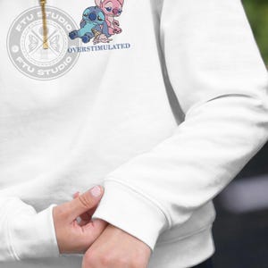 Może przedstawiać: Biała bluza z kr&oacute;tkim zamkiem błyskawicznym, ozdobiona grafiką przedstawiającą dwie niebiesko-r&oacute;żowe postacie z kresk&oacute;wek siedzące obok upuszczonego rożka z lodami. Pod postaciami znajduje się niebieski napis OVERSTIMULATED.
