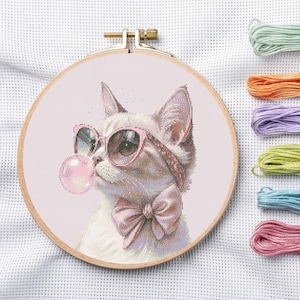 May include: Cross-stitch hoop featuring a white cat wearing pink sunglasses and a bow tie, blowing a bubblegum bubble. The cat is stitched on a light pink background. Embroidery floss in various colors is visible to the right.
