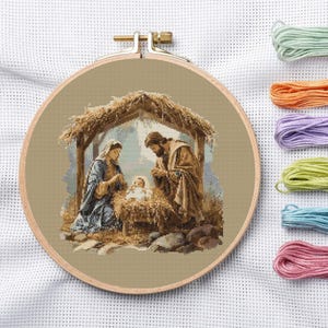 May include: A circular wooden embroidery hoop containing a cross-stitch pattern of a nativity scene with a figure in a blue robe and a figure in a brown robe standing over a baby in a manger under a thatched roof, with six skeins of thread.