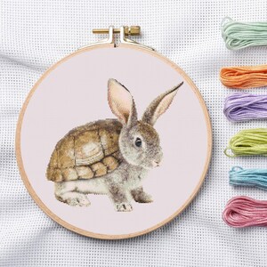 May include: Cross-stitch embroidery hoop featuring a whimsical design of a rabbit with a turtle shell. The artwork is in shades of gray, brown, and pink, with colorful embroidery floss in green, orange, purple, blue, and pink. The wooden hoop is secured with a brass clasp.