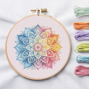 May include: A colorful cross-stitch mandala design in a wooden embroidery hoop. The mandala features a gradient of colors, including blue, yellow, orange, and pink. Several skeins of embroidery floss in matching colors are arranged to the right of the hoop.