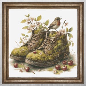 Op de afbeelding: Een ingelijst kunstwerk toont mosbedekte laarzen met bruine veters, versierd met kersenbloesems en kersen. Een kleine vogel zit op de laarzen. Het kunstwerk heeft een rustiek, natuurlijk thema.