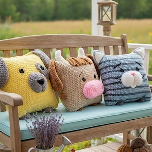 May include: Three handmade crochet animal pillows: a yellow dog, a brown cow, and a blue striped cat. The dog has gray ears and nose, the cow has a pink nose, and the cat has white ears and a white muzzle. They are sitting on a wooden bench.