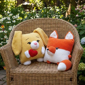 May include: Two handmade crochet pillows: a yellow bunny holding a red heart and an orange and white fox. The pillows are square-shaped and sit on a wicker chair in a garden setting. The bunny pillow has a pink nose.