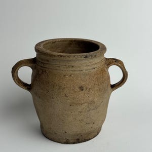 May include: An aged, earthenware jar with two loop handles. The jar has a wide mouth and a rounded form. The exterior is a mottled brown with a textured surface, indicating age and use. The jar is likely for decorative or functional use.