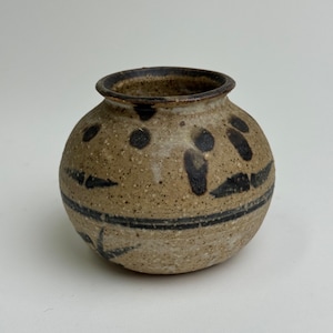 May include: A small, round ceramic vase with a speckled tan body and a dark brown rim. The vase features black, abstract markings and a horizontal band. The design is simple and rustic.