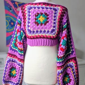 May include: A colourful, cropped crochet sweater in shades of purple, pink, teal, and yellow. The square pattern design features a central motif and balloon sleeves. The sweater is displayed on a mannequin.