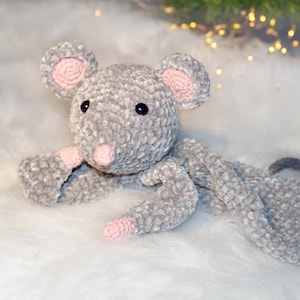 Könnte beinhalten: Eine handgefertigte graue Häkelmaus mit rosa Akzenten an Ohren, Nase und Füßen. Das Plüschtier hat schwarze Knopfaugen und einen grauen Schwanz. Die Maus liegt auf einer weißen, flauschigen Oberfläche.