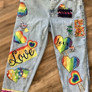 May include: Light blue denim jeans featuring various colorful patches including rainbow hearts, floral patterns, and a rectangular patch with the word Love in black script. The jeans have a frayed hem with a striped trim at the bottom edge.