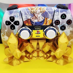 Puede incluir: Mando de videojuegos personalizado con un gráfico de un personaje de Dragon Ball Z. El mando tiene una base dorada con detalles de cristal. El texto "OPELLOCUSTOMS" es visible en la parte superior.