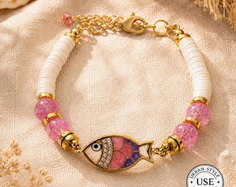 May include: Gold-toned bracelet featuring a fish charm with pink, purple, and white enamel. The bracelet includes white disc beads, pink crystal beads, and a gold chain. The charm has a rhinestone eye and is labeled with "Urban Style Essentials".