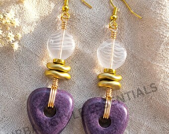 May include: Dangle earrings featuring a purple heart-shaped stone bead, two gold-toned flat circular spacers, and a translucent white glass bead with swirling patterns. The components are connected by gold-toned wire and attached to gold-toned hooks.