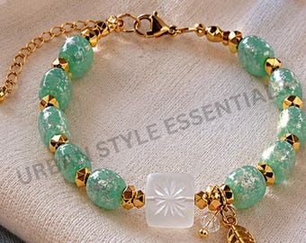 May include: A bracelet featuring mint green beads with silver flecks, interspersed with small gold-toned metal beads. A central square white bead with a starburst engraving is present, along with a gold-toned leaf charm and a lobster claw clasp.