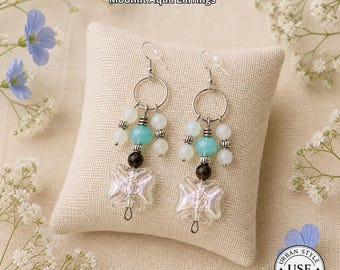 May include: A pair of Moonlit Aqua Earrings with silver-tone hoops and ear wires. The earrings feature light blue, white, and black beads, with a clear, butterfly-shaped charm at the bottom. The text "URBAN STYLE ESSENTIALS" is at the top.