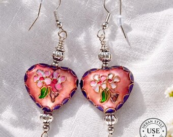 May include: A pair of heart-shaped earrings with a pink and purple floral design. The earrings have silver-colored hooks and decorative beads. The heart-shaped pendants feature a pink background with white and pink flowers and green leaves.