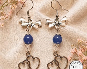 May include: A pair of silver-toned earrings with a bow design at the top, a blue bead, clear beads, and two intertwined hearts at the bottom. The earrings are displayed on a beige fabric.