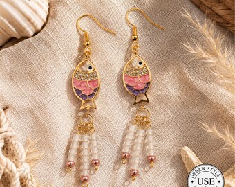 May include: A pair of gold-colored fish-shaped earrings. The fish are painted with pink, purple, and white enamel and adorned with small crystals. Each earring has a dangling strand of white beads with pink accents.
