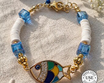 May include: Gold-toned bracelet featuring a colorful fish pendant with a blue eye and crystal accents. The bracelet includes blue cube beads, white disc beads, and gold-colored chain links. The bracelet is a stylish accessory.