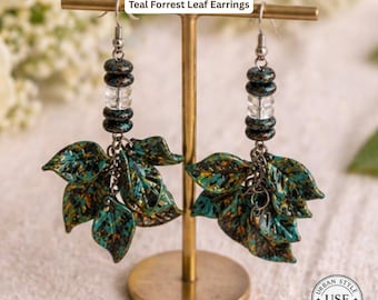 May include: Teal Forrest Leaf Earrings. These earrings feature leaf-shaped pendants with teal, brown, and yellow accents. The earrings are suspended from a silver chain and are displayed on a gold-colored stand. The text "Urban Style Essentials" is at the top.