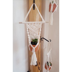 May include: A white macrame plant hanger with a terracotta pot holding a green succulent. The macrame design features intricate knots and a tassel. A wooden dowel supports the hanger, which is mounted on a wooden post.