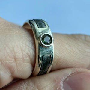 May include: A silver ring with a dark green gemstone set in a circular bezel. The band features rectangular inlays of a dark, textured material. The ring is shown on a finger, with a light blue background.