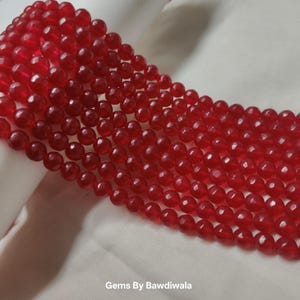 May include: Close-up of several strands of faceted, translucent red beads. The beads are round and have a glossy finish. The image is set against a white background. The text "Gems By Bawdiwala" is at the bottom.