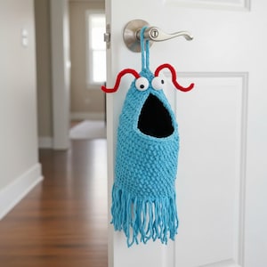 May include: A whimsical, handmade blue crochet door organizer shaped like a monster, complete with googly eyes and red antennae. The open mouth design features a black interior and a fringe bottom. It hangs from a silver door handle.
