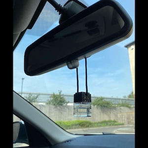 May include: A black and gold car air freshener hanging from the rear view mirror of a car. The air freshener is a small square bottle with a clear liquid inside.