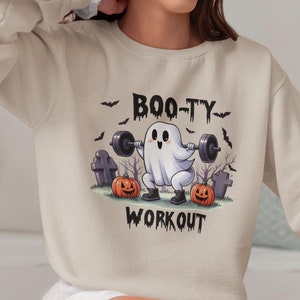 Puede incluir: Una sudadera de color beige claro con un gráfico de un fantasma levantando pesas con el texto "BOO-TY WORKOUT" impreso en ella.