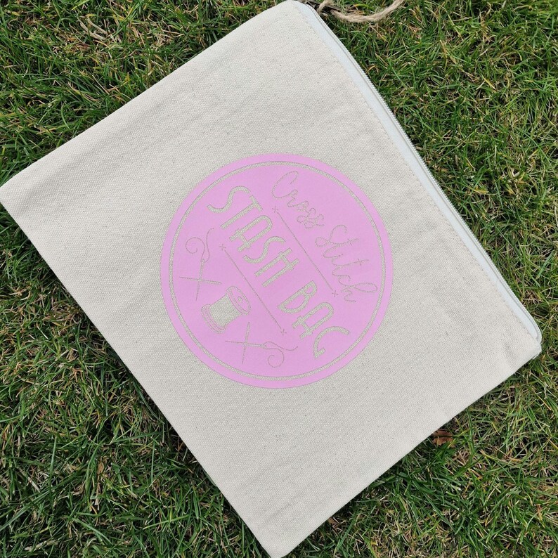 May include: A beige canvas zippered pouch with a pink circle that says "Cross Stitch Stash Bag" in white text. The circle also has a stylized image of scissors and a spool of thread.