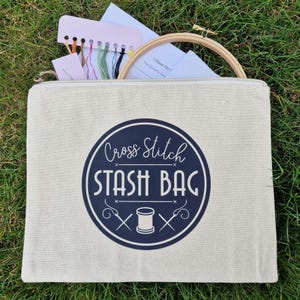 May include: A white canvas zippered pouch with a blue circle logo that says "Cross Stitch Stash Bag" with a spool of thread and two needles crossed inside the circle.