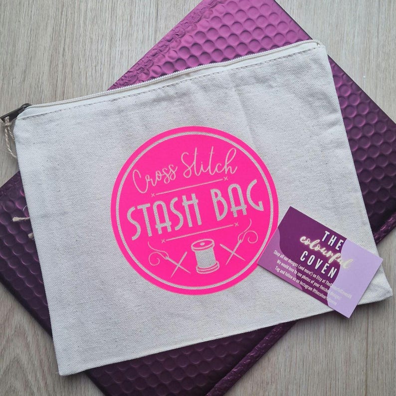 May include: A white canvas zippered pouch with a pink circle logo that says "Cross Stitch Stash Bag" with a pink needle and thread design. The pouch is sitting on a purple bubble mailer with a business card that says "The Colourful Coven" in pink and white.
