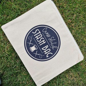 May include: A white canvas zippered pouch with a navy blue circle logo that says "Cross Stitch Stash Bag" with a needle, thread, and scissors graphic.