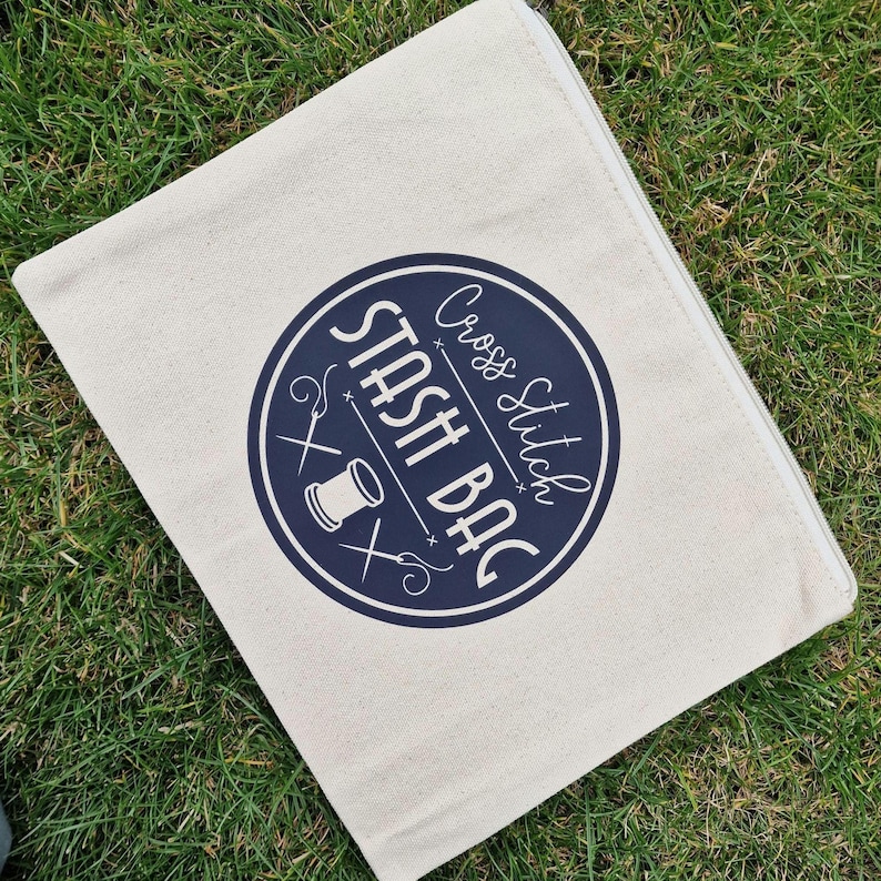 May include: A white canvas zippered pouch with a navy blue circle logo that says "Cross Stitch Stash Bag" with a needle, thread, and scissors graphic.