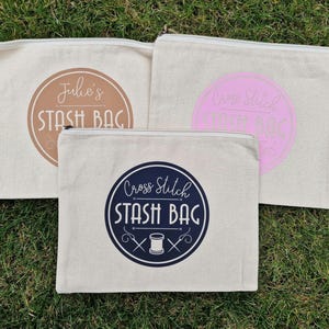 May include: Three white canvas zippered pouches with different designs. The first pouch has a brown circle with the text "Julie's Stash Bag". The second pouch has a pink circle with the text "Cross Stitch Stash Bag". The third pouch has a blue circle with the text "Cross Stitch Stash Bag" and a needle and thread graphic.