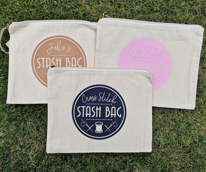 May include: Three white canvas zippered pouches with different designs. The first pouch has a brown circle with the text "Julie's Stash Bag". The second pouch has a pink circle with the text "Cross Stitch Stash Bag". The third pouch has a blue circle with the text "Cross Stitch Stash Bag" and a needle and thread graphic.