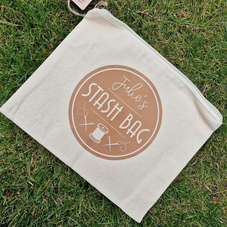 May include: A white canvas zippered pouch with a brown circle logo that says "Julie's Stash Bag" with a stylized image of a spool of thread and needles.