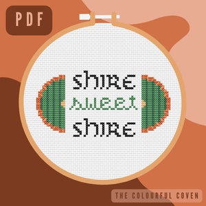 May include: A white embroidery hoop with a green and orange cross-stitch design of a hobbit door and the text "shire sweet shire".