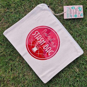May include: A white canvas zippered pouch with a red circle design that says "Natasha's Stash Bag" in white text. The circle has a red glitter effect and a small image of a needle and thread.