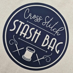 May include: A navy blue circle with white text that reads "Cross Stitch Stash Bag". The circle has two white needle and thread designs and a white spool of thread.
