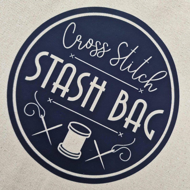 May include: A navy blue circle with white text that reads "Cross Stitch Stash Bag". The circle has two white needle and thread designs and a white spool of thread.