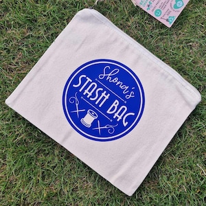 May include: A white canvas zippered pouch with a blue circle design. The circle has the text "Shona's Stash Bag" in white lettering and a white sewing needle and thread graphic.