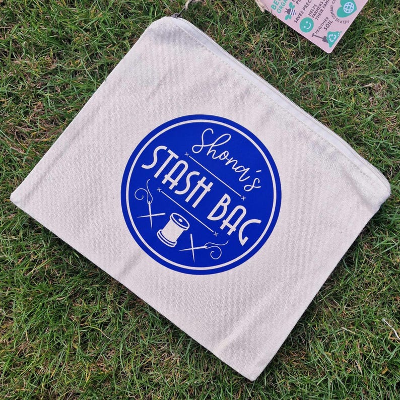 May include: A white canvas zippered pouch with a blue circle design. The circle has the text "Shona's Stash Bag" in white lettering and a white sewing needle and thread graphic.